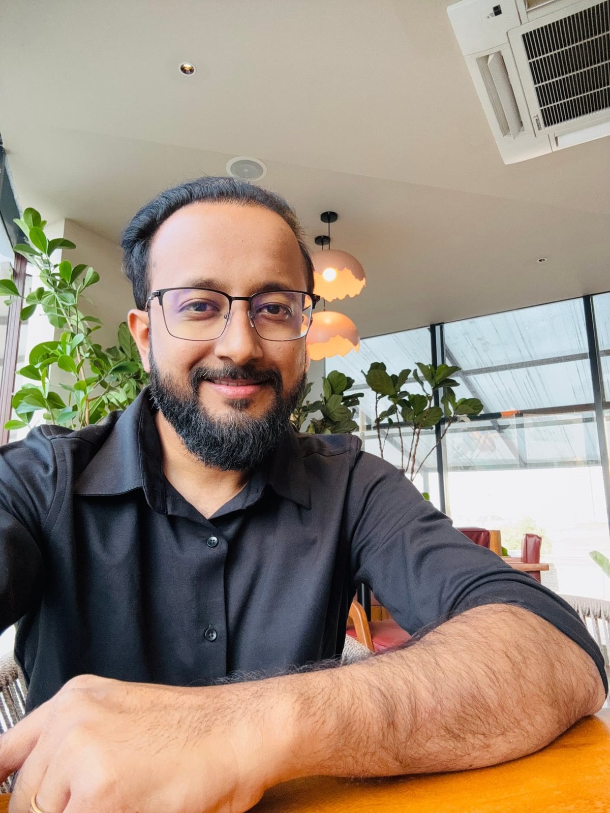 Akshay Katkade, smiling, seated at a wooden table in a bright café, wearing glasses and a black shirt.